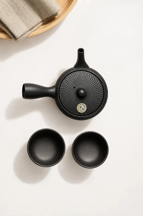 A black cast-iron teapot with a lid featuring a sunburst pattern and a small gold emblem, shown alongside two matching black cups on a white background.