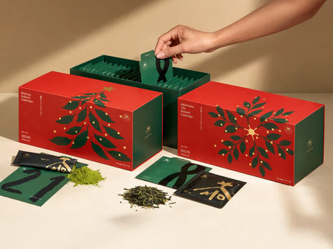 A hand reaches into a red and green gift box set on a beige surface, with two matching decorative boxes, small cards, loose green tea, and a few tea bags arranged nearby.