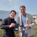 Oishi farmer and Nio Teas' CEO standing outdoors in a green tea plantation holding green tea  pouches with white labels under a blue sky.