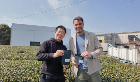 Oliver Staerkle and Oishi farmer standing outdoors in a tea plantation holding black green tea bags, with industrial buildings and a blue sky in the background.